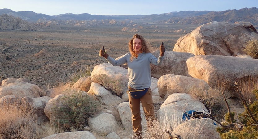 a veteran smiles and gives two thumbs up to the camera while standing on a rock formation in front of the vast desert landscape of joshua tree